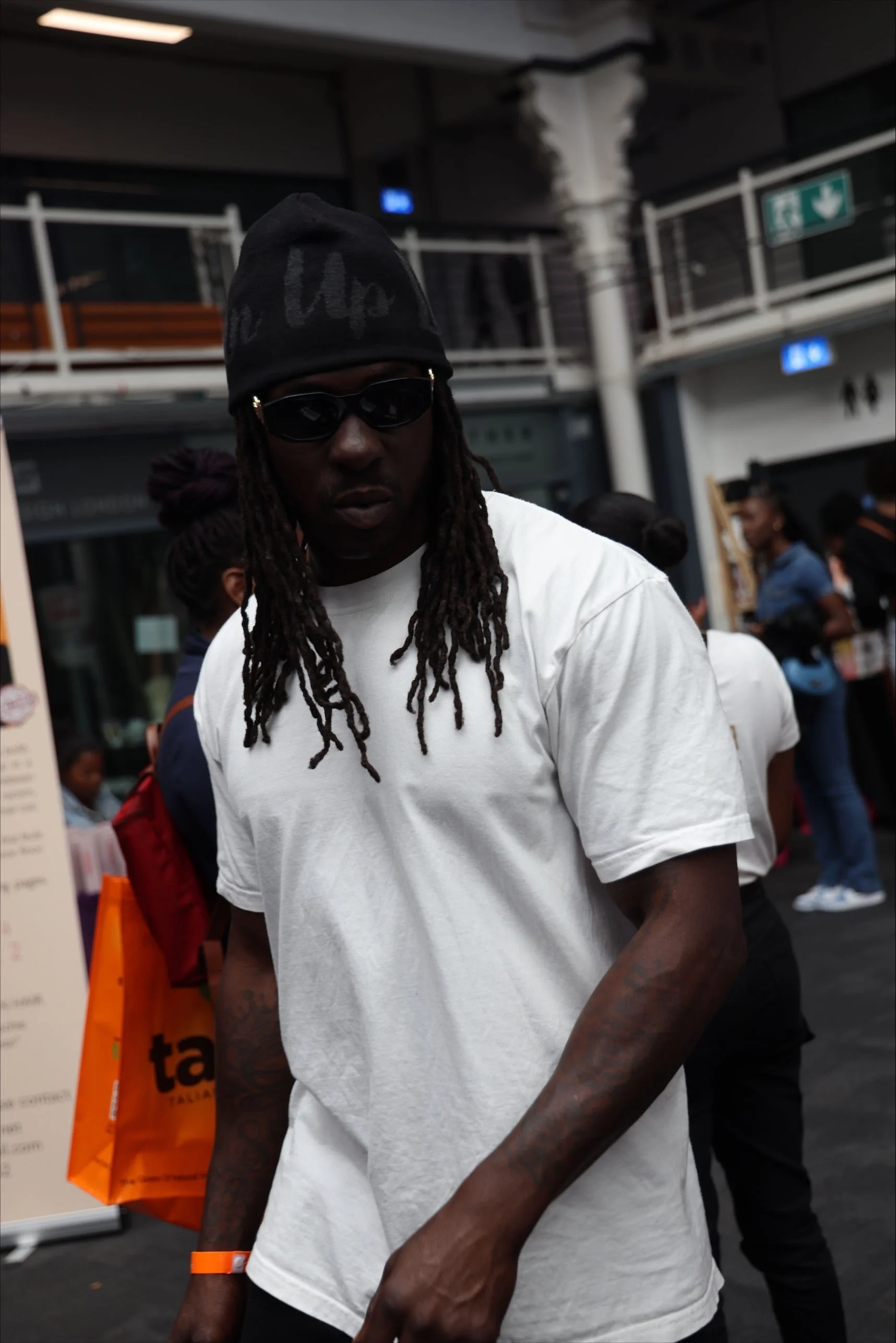A man with dreadlocks wearing black sunglasses, a black beanie, and a white t-shirt, standing in a busy indoor location with other people around.