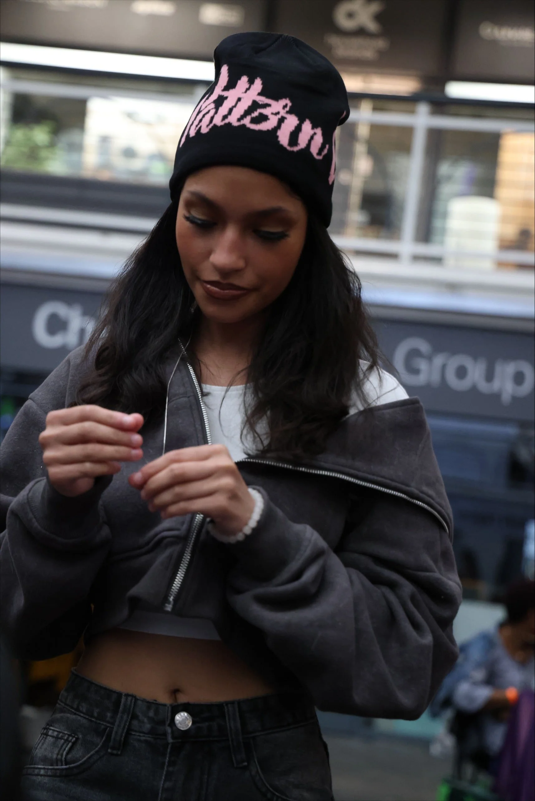 A young woman with long dark hair wearing a black beanie with pink lettering, a gray zip-up hoodie, and dark jeans, looking down and adjusting her hoodie in an indoor public space.