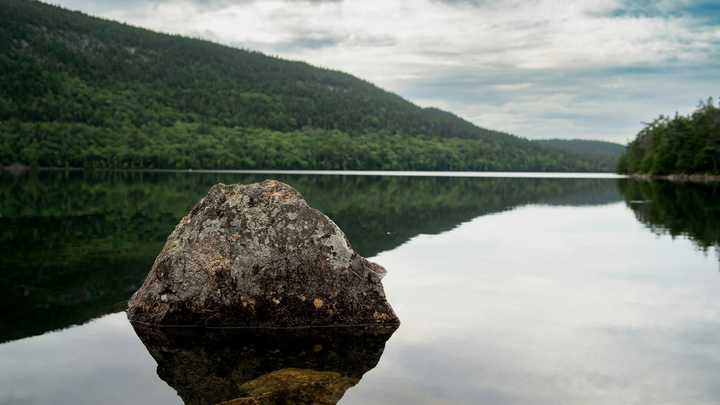 A large rock in a calm lake with reflections of the surrounding green forested hills and cloudy sky.