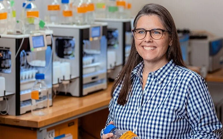 A woman with glasses wearing a blue and white checkered shirt standing in a laboratory with scientific equipment and test tubes in the background.