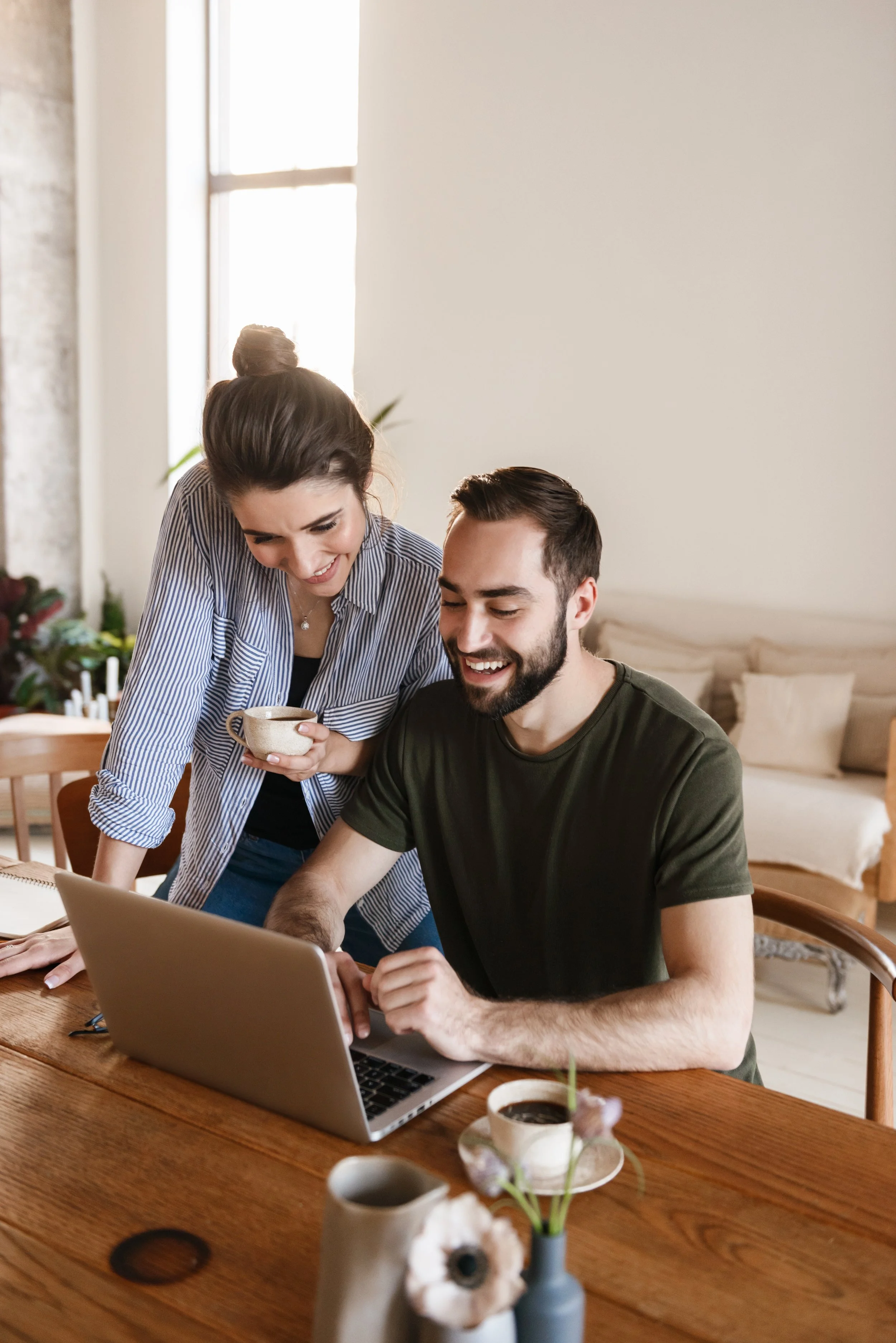 A woman and a man are looking at a laptop together in a bright room, smiling and sharing a moment of laughter. The woman is holding a mug, and there are flowers and cups on the wooden table.