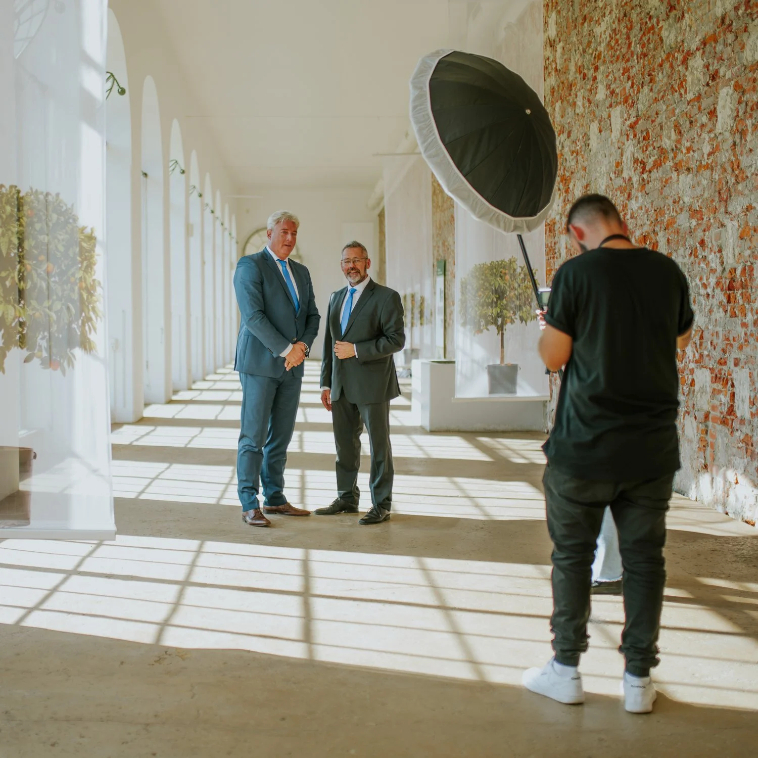 Zwei Männer in Anzügen werden in einem hellen Raum mit Sonnenlicht durch große Fenster fotografiert, während ein Fotograf mit Lichtausrüstung Fotos macht.