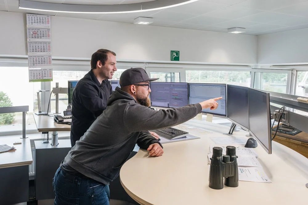Zwei Männer diskutieren vor mehreren Computermonitoren in einem Büro, eines zeigt eine Tabelle oder einen Bericht, während sie auf den Bildschirm zeigen.