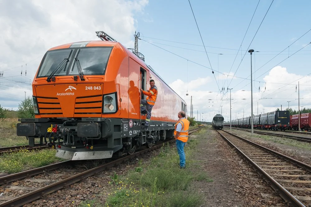 Zwei Arbeiter in Sicherheitswesten arbeiten an einer orangefarbenen elektrischen Lokomotive auf Gleisen, mit weiteren Zügen im Hintergrund.
