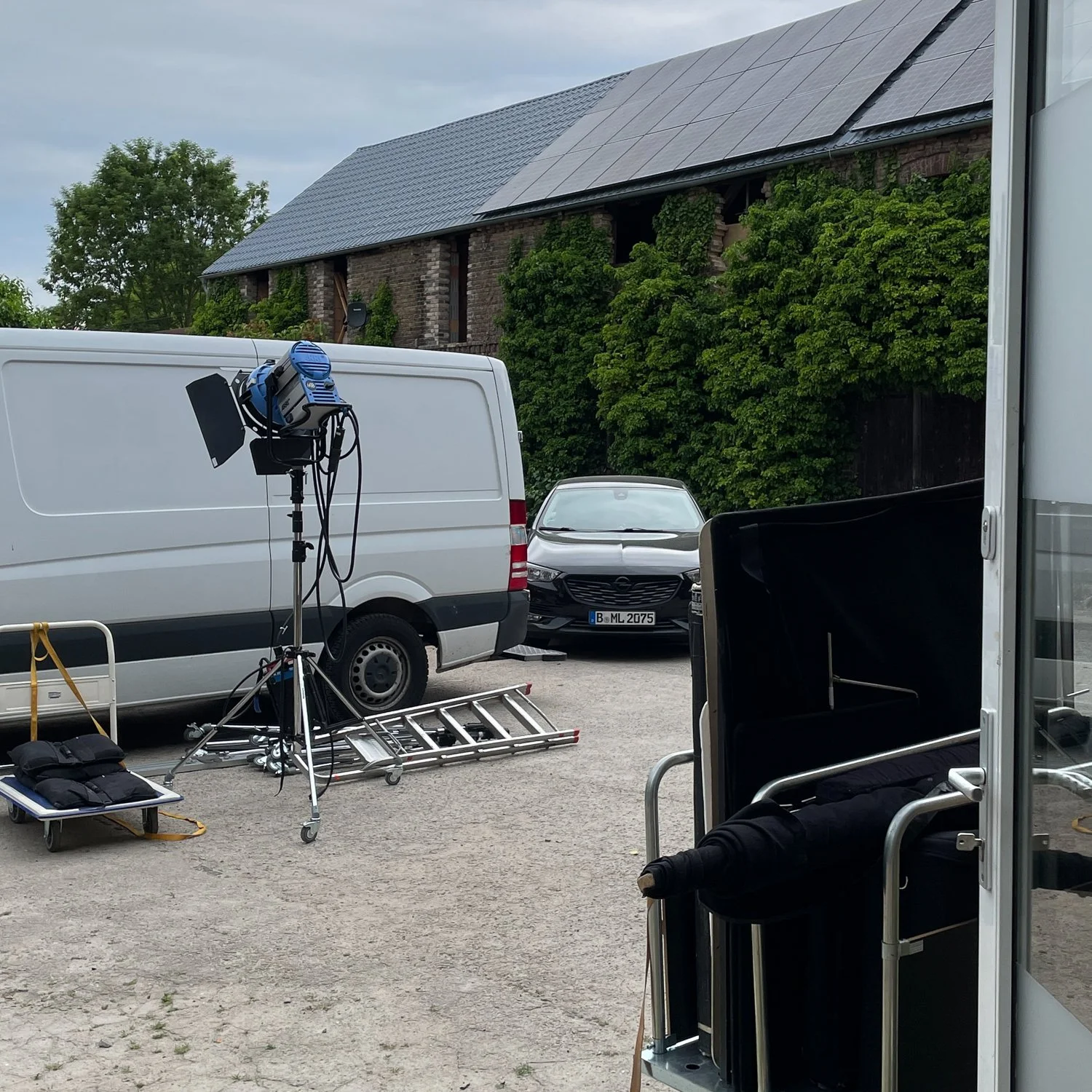 Filmset oder Fotoshooting-Ausrüstung im Freien mit Lichttechnik, Kamera, Leiter und Transportkoffer, in der Nähe eines Hauses mit Solardach und bewachsenen Wänden, mit Fahrzeugen im Hintergrund.