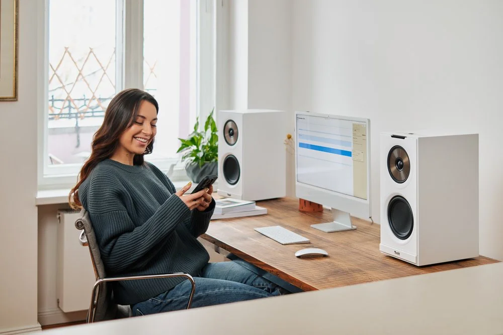 Junge Frau sitzt an einem Schreibtisch, lächelt und schaut auf ihr Smartphone, mit einem Computer und Lautsprechern im Hintergrund.