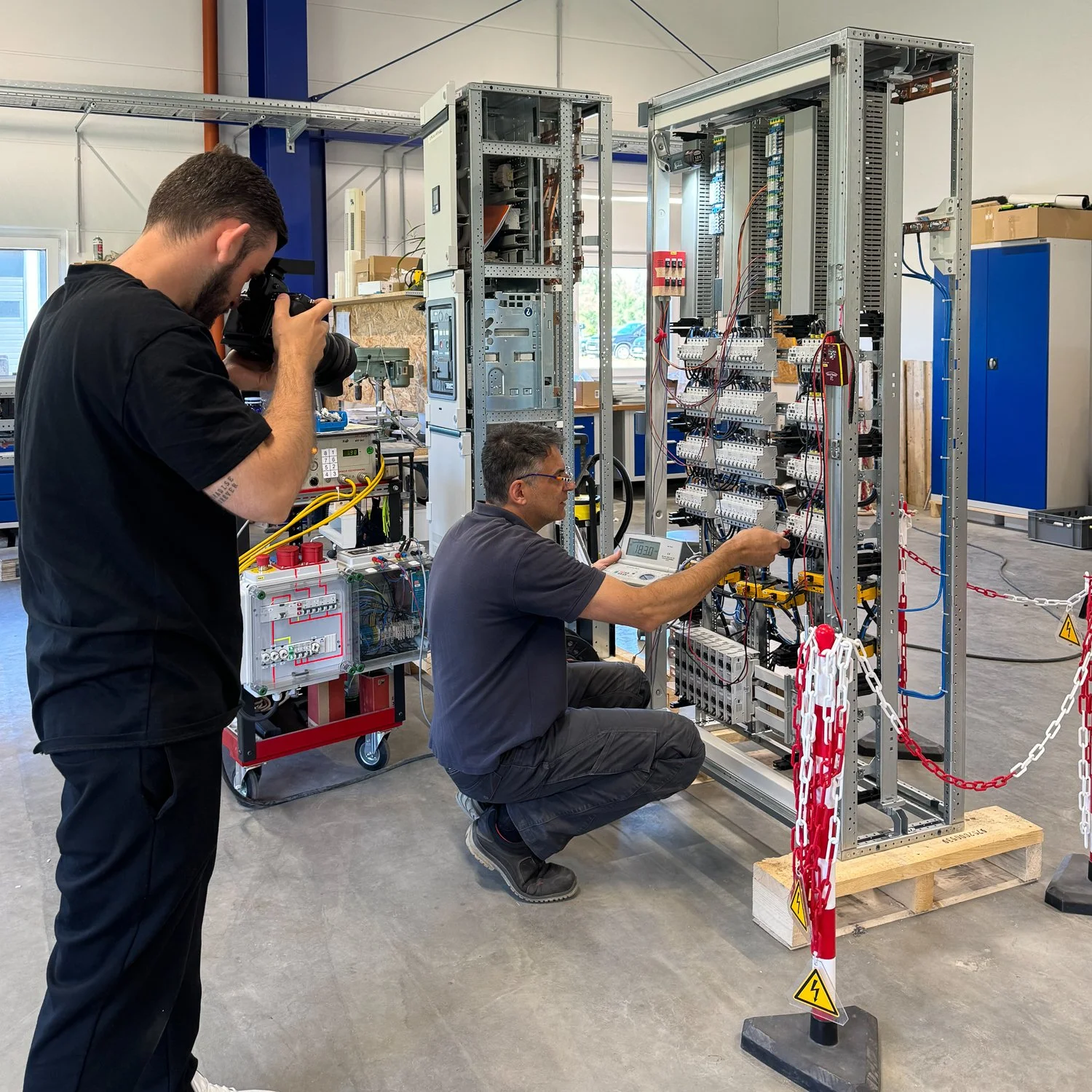 Zwei Techniker arbeiten an einem offenen elektrischen Steuerungskasten in einer Werkstatt, während andere eine Kamera für eine Aufnahme nutzen.