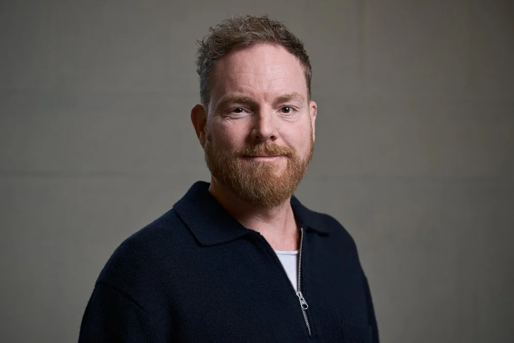 Ein Mann mit einem roten Bart, kurzen braunen Haaren, trägt ein dunkles Zip-Hemd vor einem neutralen grauen Hintergrund.