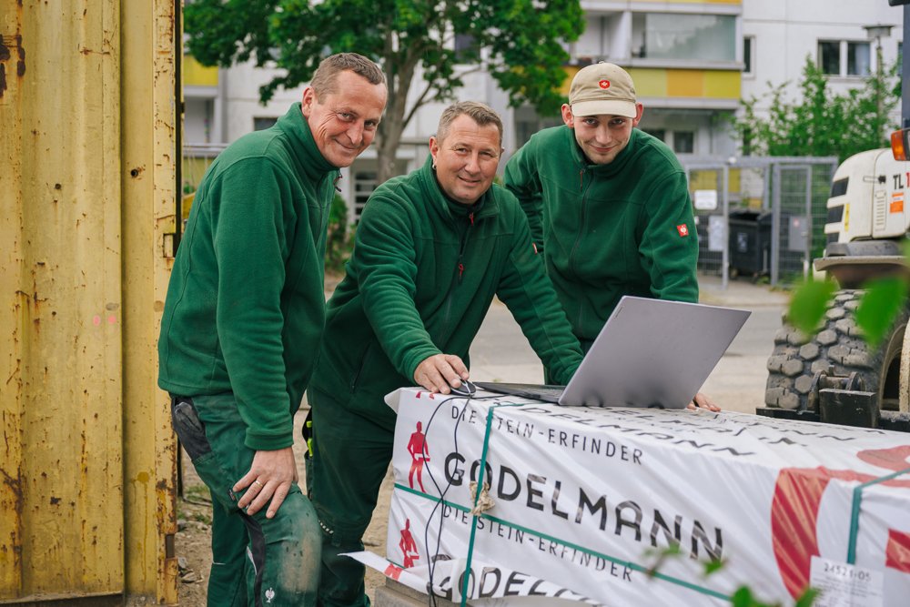 Drei Männer in grünen Jacken vor einem Laptop, die an einem Tisch mit einem Banner arbeiten, im Hintergrund eine Baustelle und eine grüne Umgebung.