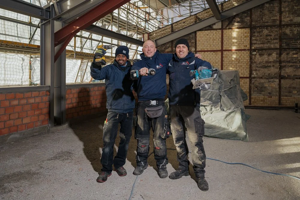 Drei Bauarbeiter in Arbeitskleidung, die in einem Bauwerk im Rohbau arbeiten, stehen zusammen und lächeln in die Kamera.