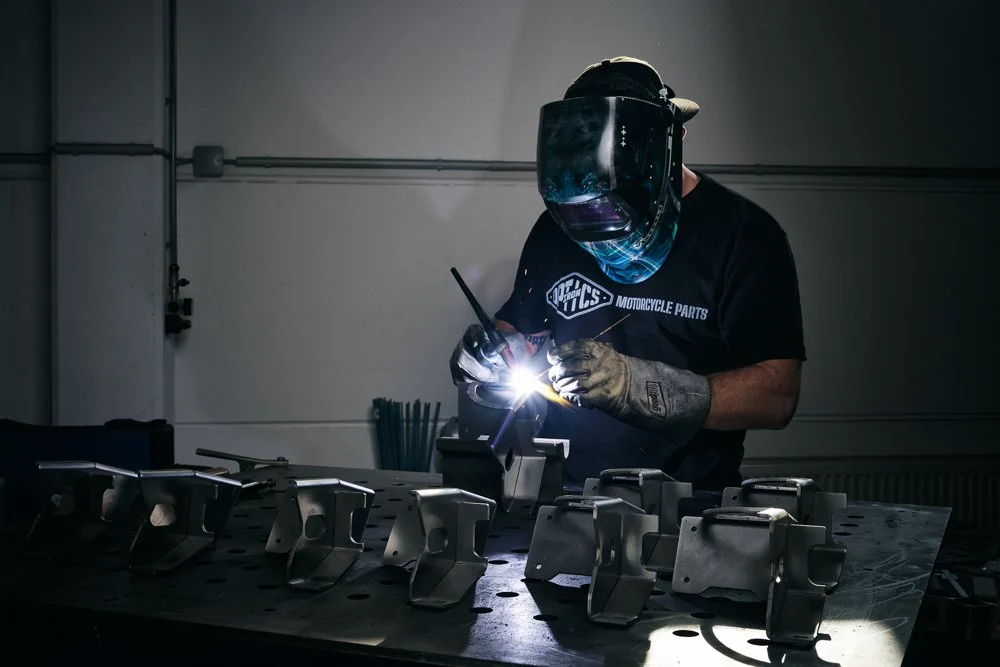 Mensch schweißt Metallteile in einer Werkstatt, trägt Schutzhelm und Handschuhe.