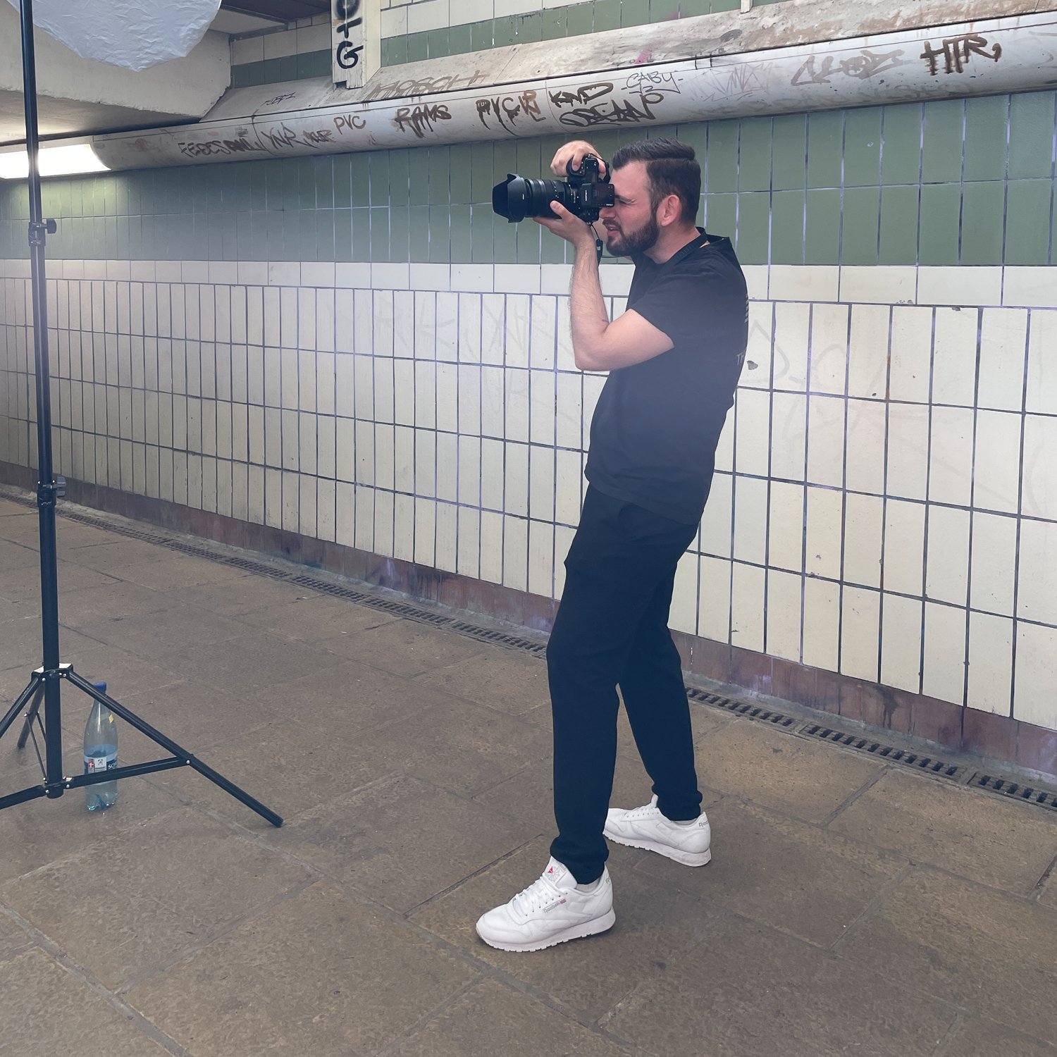 Ein Mann mit dunklem Haar und Bart, der eine Kamera hält und fotografiert, steht in einem öffentlichen Tunnel oder U-Bahnhof, neben einer Wand mit weißen und grünen Fliesen. Auf dem Boden steht eine Wasserflasche, und es gibt ein Stativ mit einer Bel