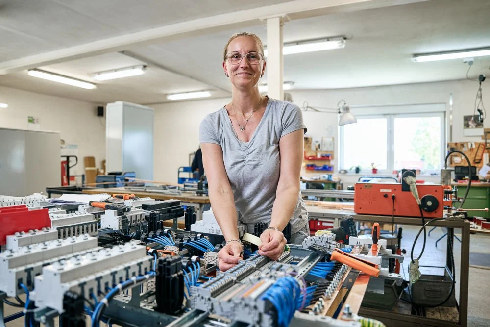 Eine Frau arbeitet in einer Werkstatt mit elektromagnetischer Verkabelung, umgeben von Werkzeugen und elektrischen Komponenten.
