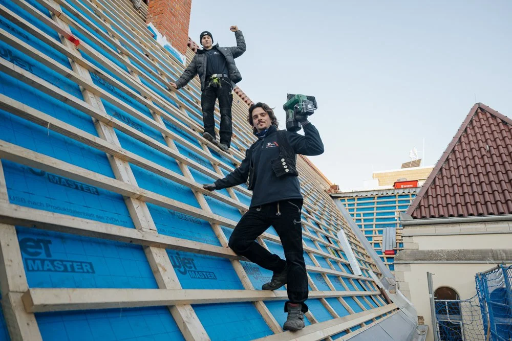Zwei Männer auf einem großen, im Bau befindlichen Dach, eines hält eine elektrische Schraubpistole, das andere zeigt mit dem Arm nach oben.