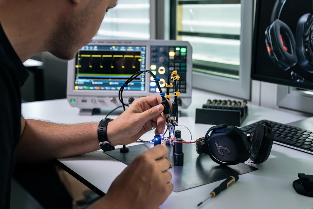 Techniker arbeitet mit Messgeräten an einem elektronischen Gerät, während im Hintergrund ein Oszilloskop und ein Monitor mit Messdaten zu sehen sind, in einem Raum mit Fenster.