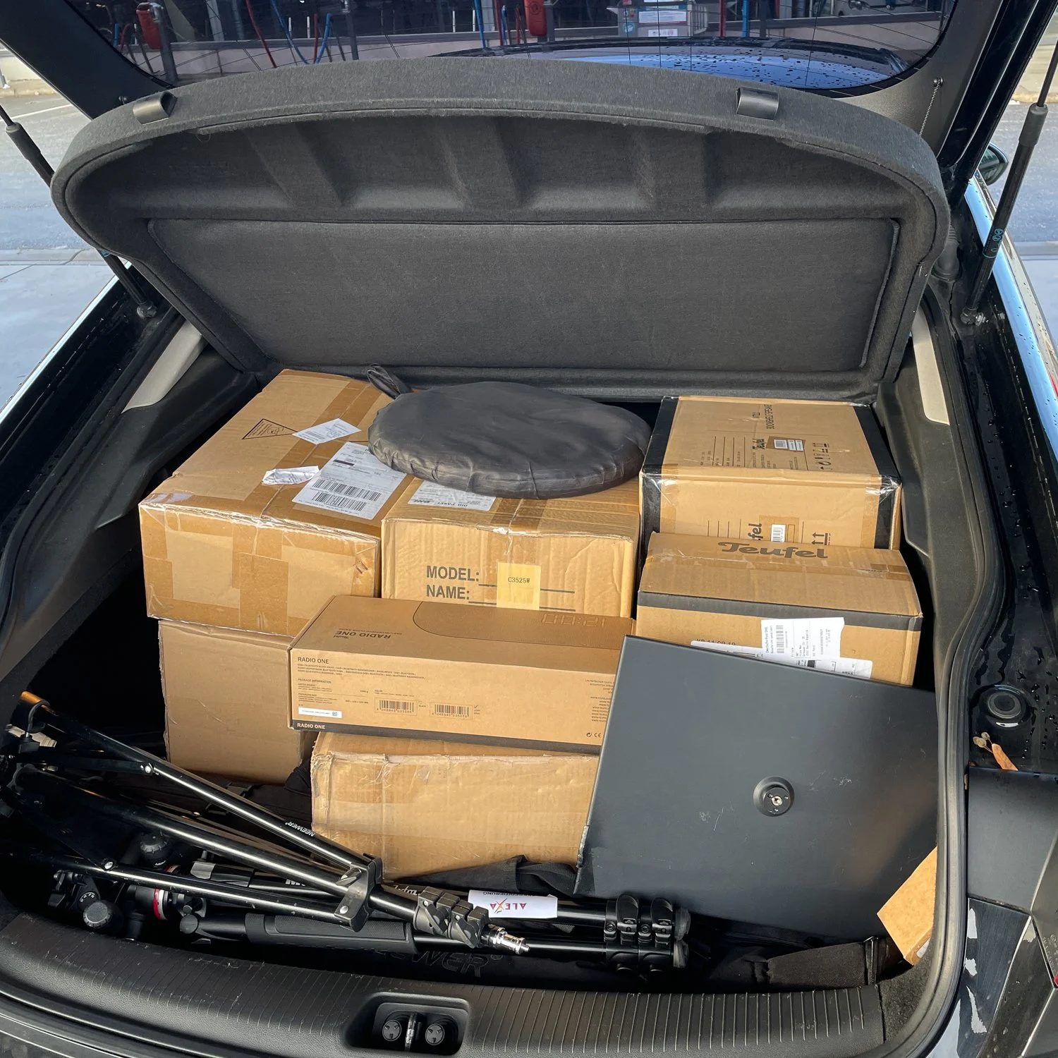 Ein Kofferraum mit mehreren Kartons, einem schwarzen sechseckigen Gegenstand und einem zusammengefalteten Stativ.