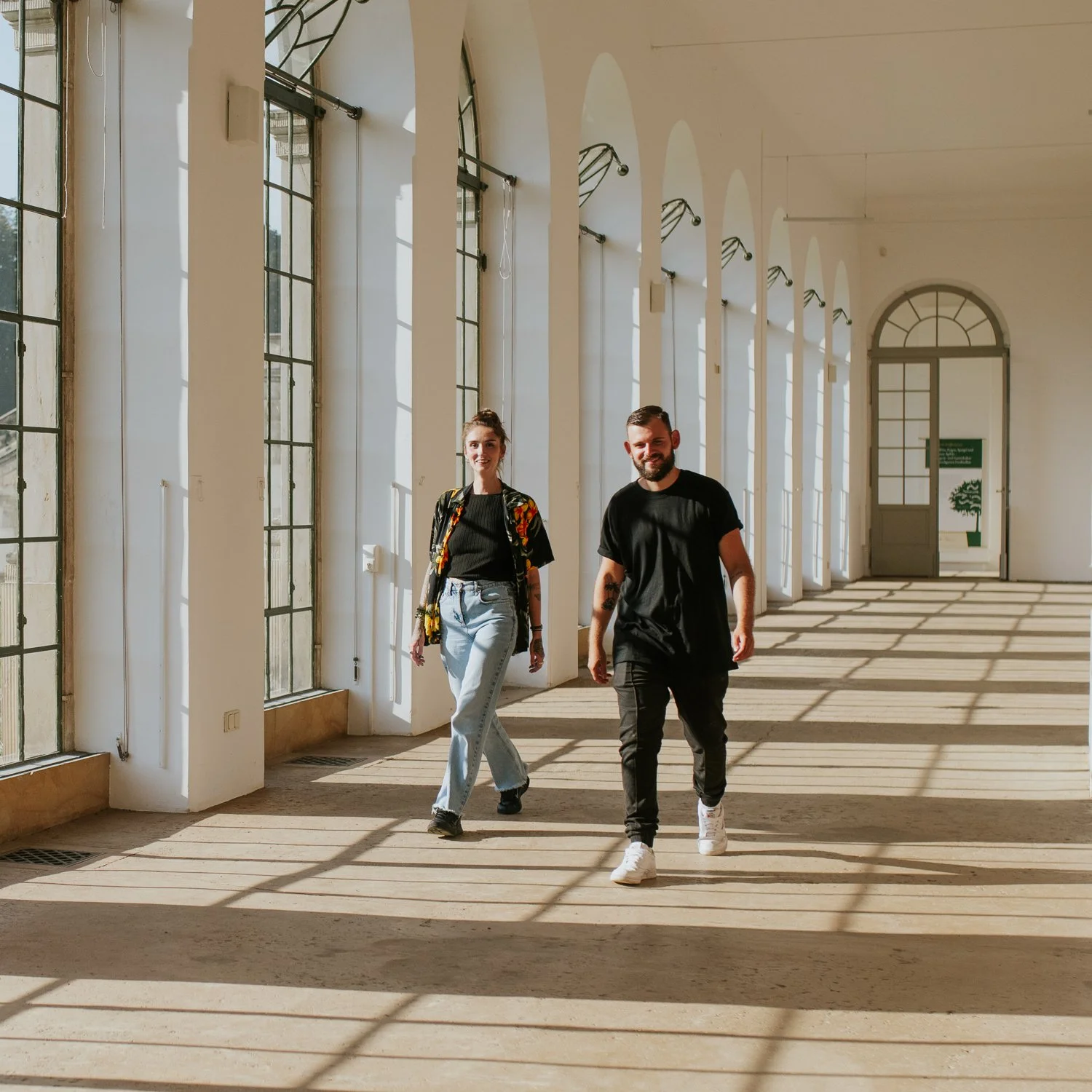 Zwei Menschen gehen durch einen lichtdurchfluteten Raum mit hohen Fenstern und Schattenmustern auf dem Boden.
