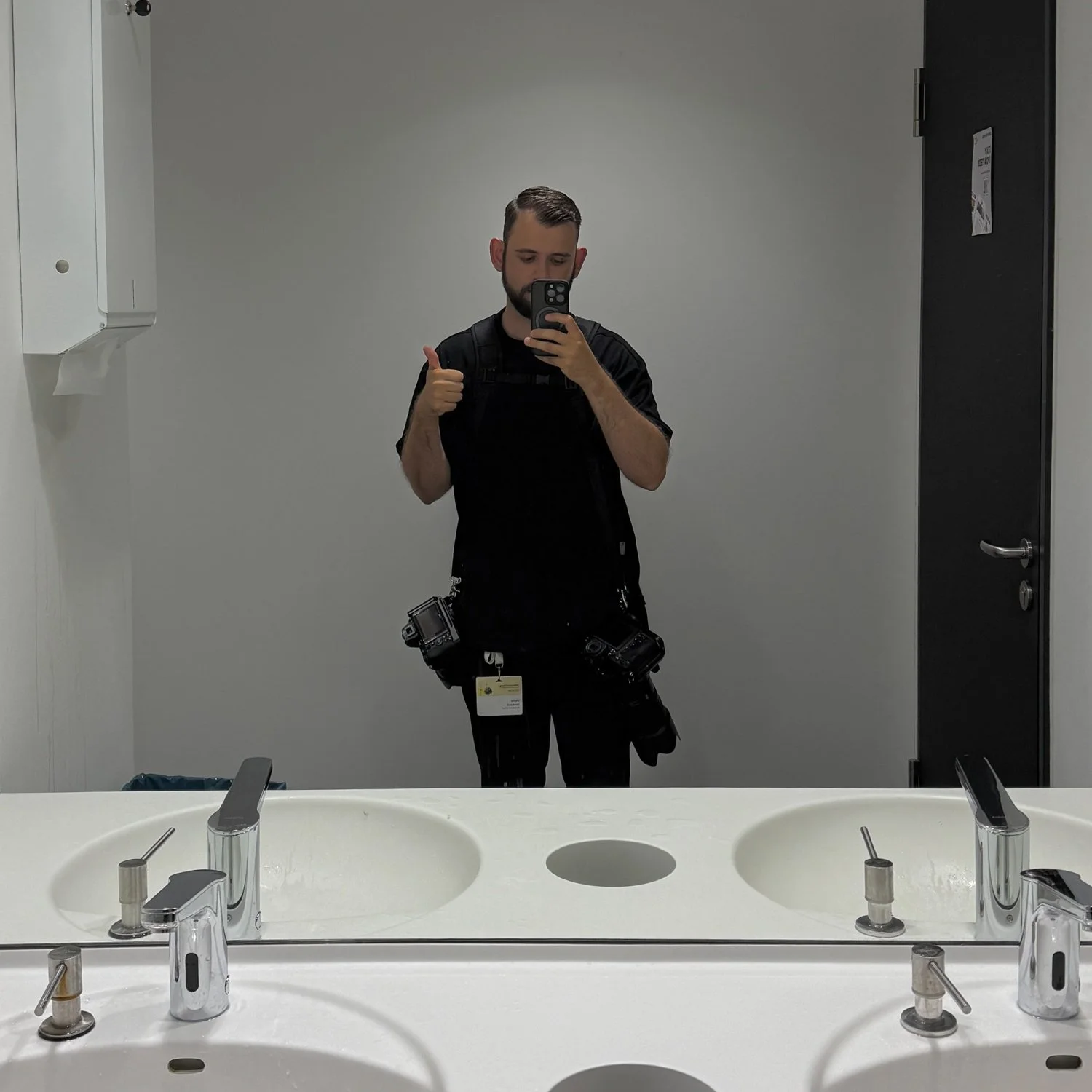 Ein Mann in einem Badezimmer macht ein Selfie im Spiegel. Er trägt schwarze Kleidung und hat eine Kameraausrüstung um die Hüften. Er zeigt mit einer Hand einen Daumen nach oben.