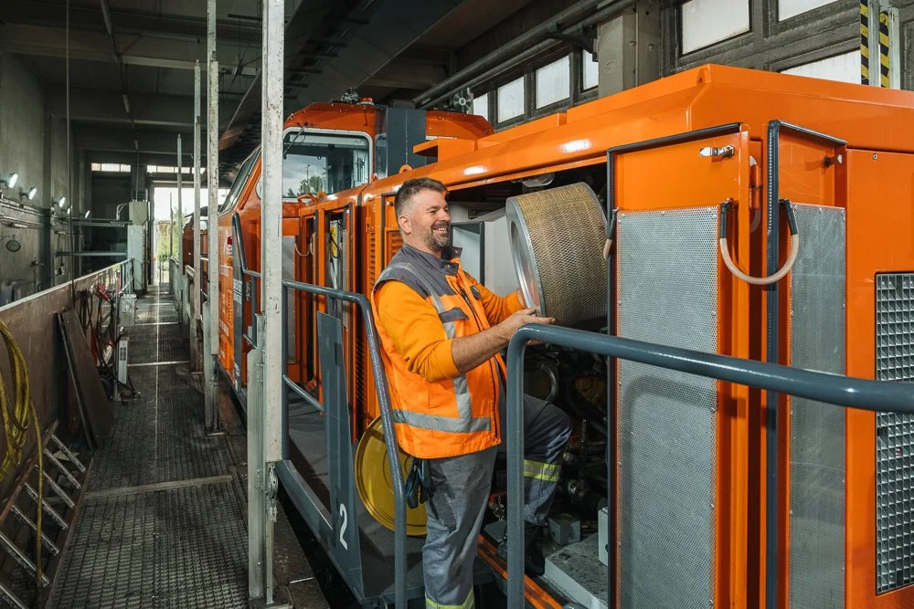 Mitarbeiter in orange Sicherheitsweste arbeitet an einer großen orangefarbigen Maschine in einer Werkshalle.
