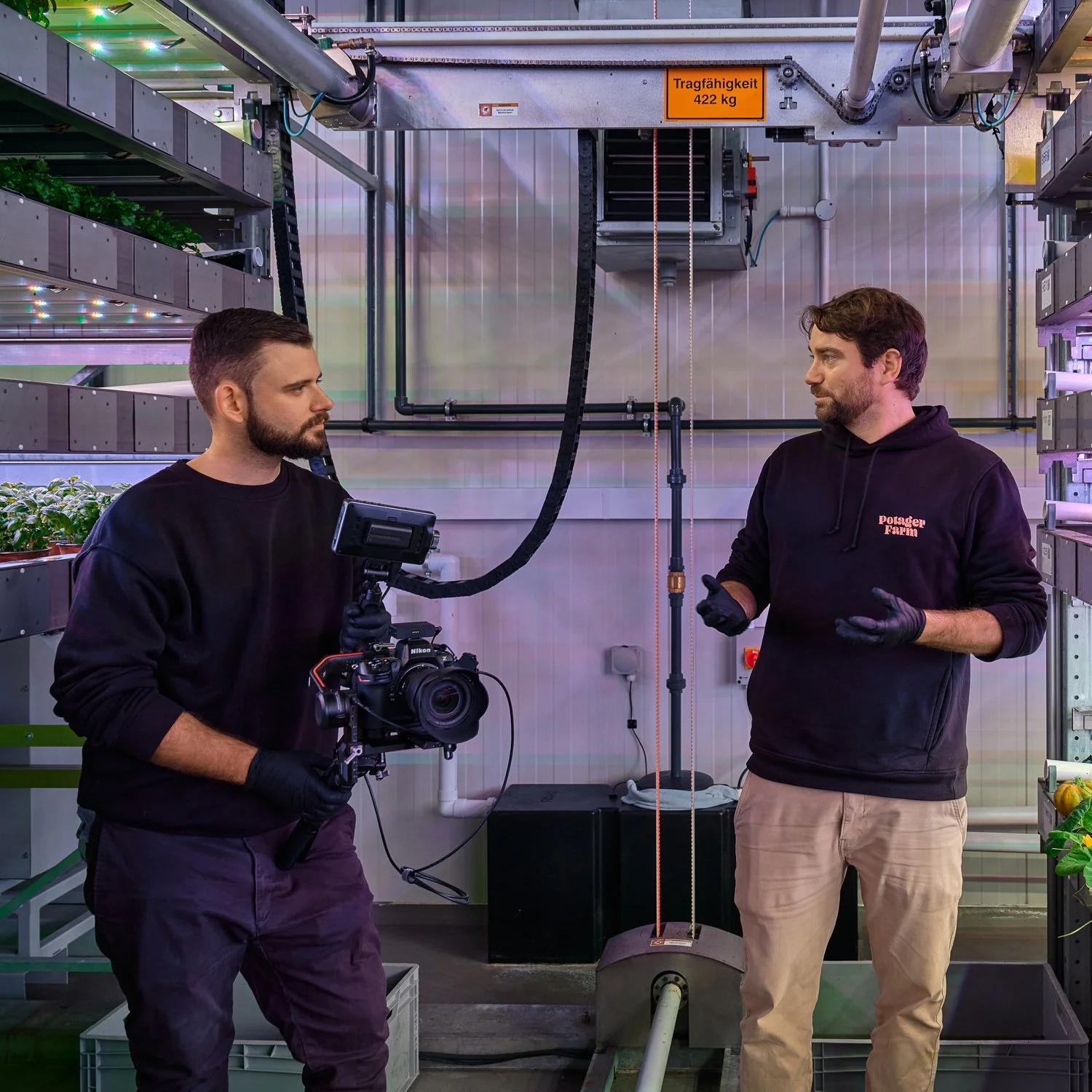 Zwei Männer, die in einer Pflanzenzuchtanlage sprechen. Der Mann auf der linken Seite hält eine Kamera, der Mann auf der rechten Seite trägt schwarze Handschuhe und ist in einem schwarzen Hoodie mit rotem Text. Im Hintergrund sind Pflanzen und techni
