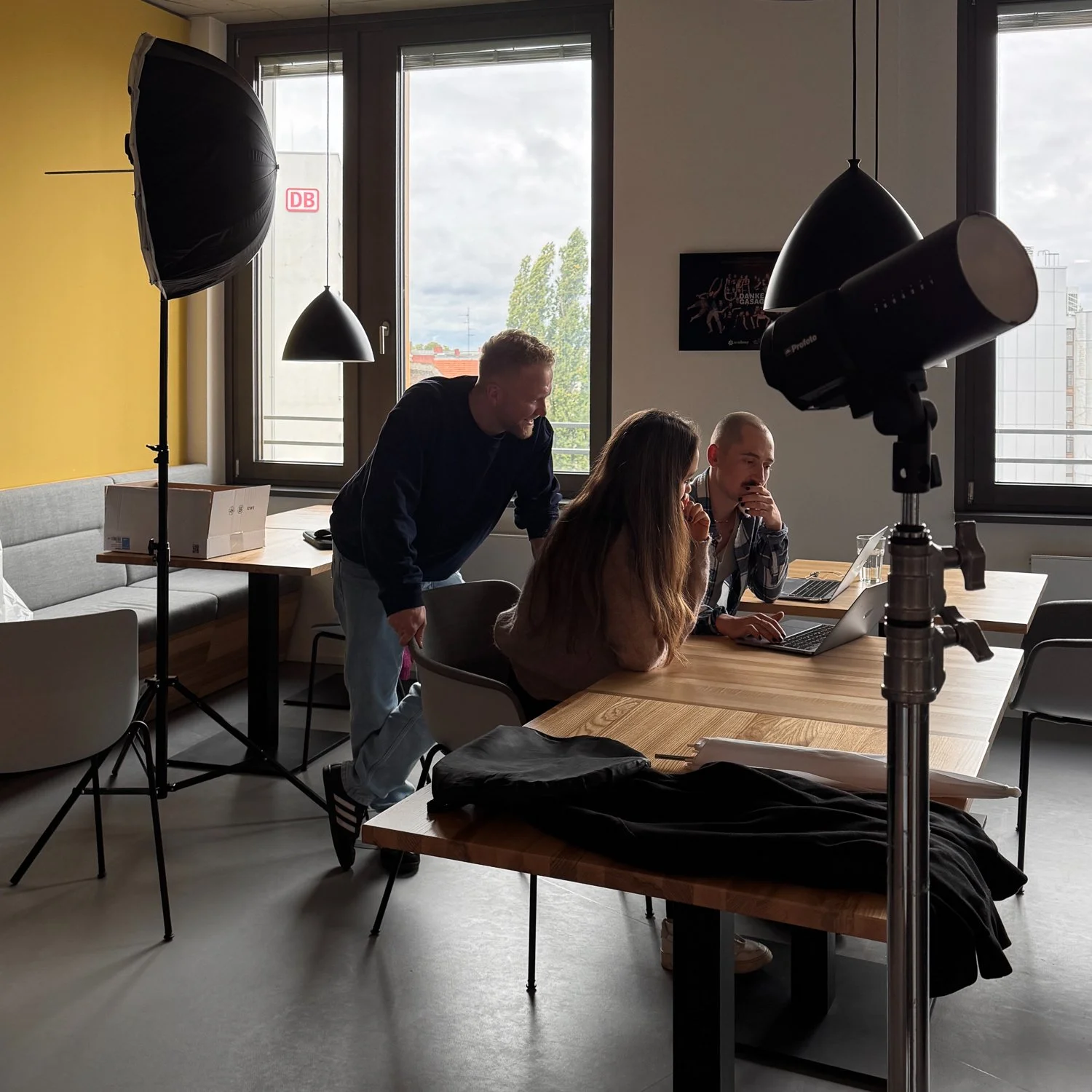 Drei Menschen arbeiten an einem Tisch in einem Raum mit großen Fenstern, während sie eine Kamera und Fotolicht für eine Fotosession vorbereiten.