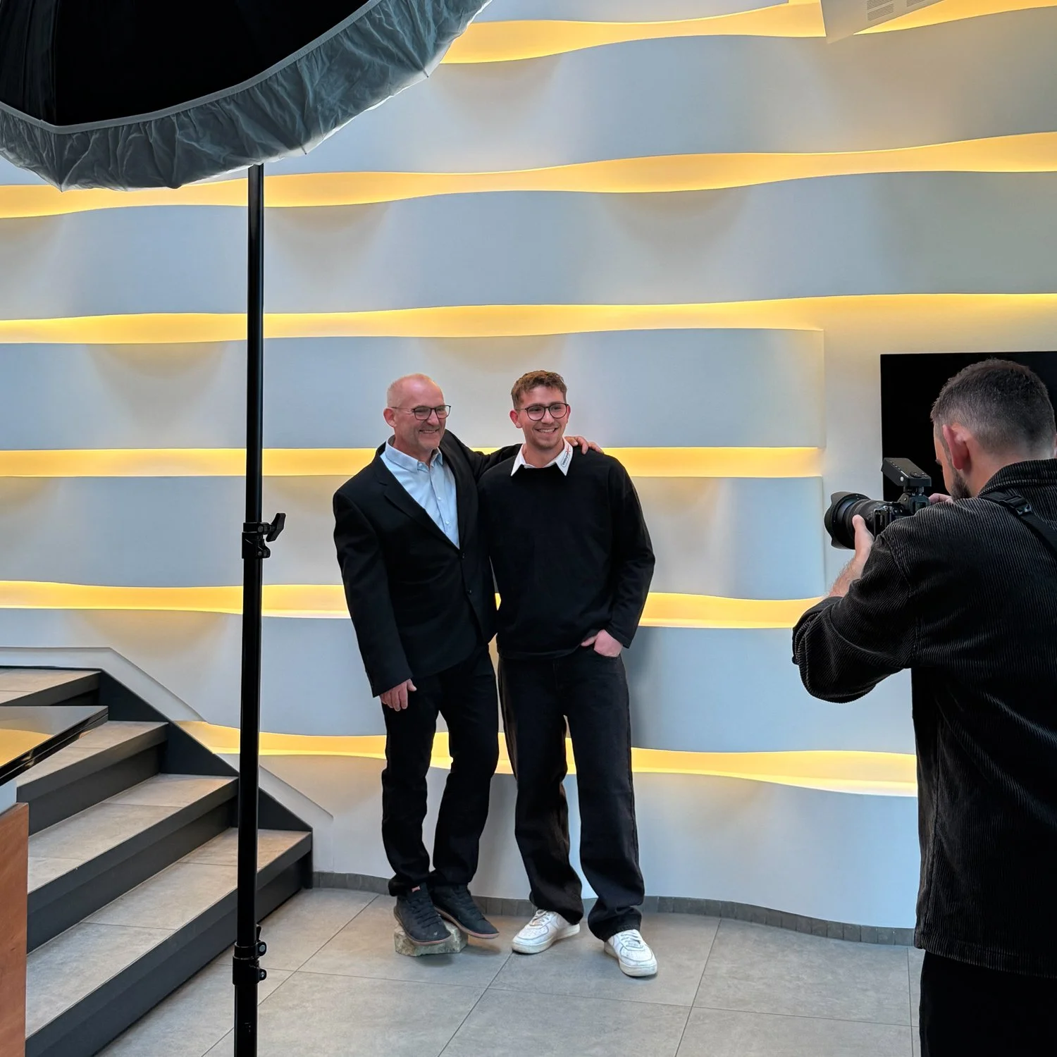 Zwei Männer posieren für ein Foto, einer mit Brille, der andere im schwarzen Pullover, im Hintergrund eine modern gestaltete Wand mit horizontalen, gelb beleuchteten Linien, im Vordergrund ein Fotograf, der das Bild aufnimmt.