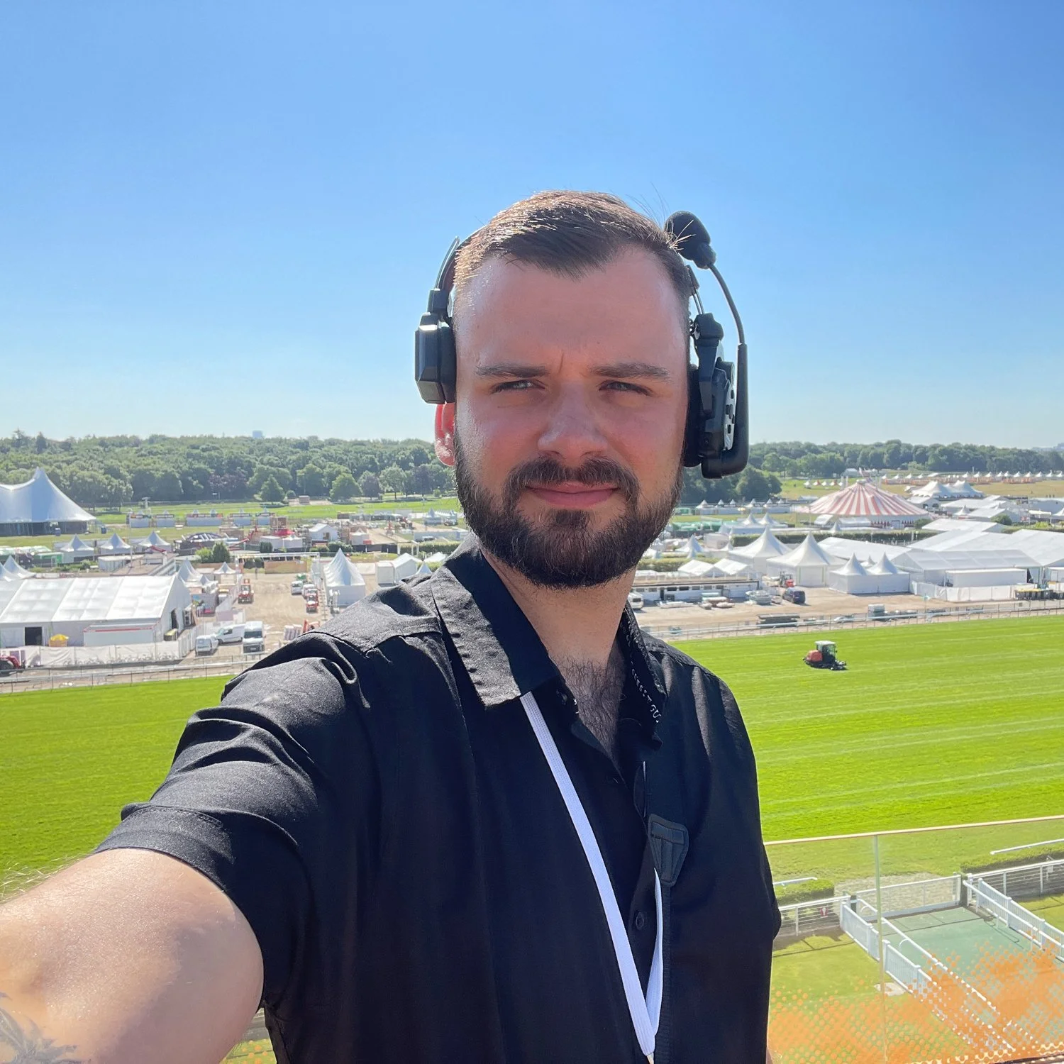 Ein Mann mit einem schwarzen Hemd trägt ein Headset und macht ein Selfie auf einem Festivalgelände mit weißen Zelten und einer grünen Wiese im Hintergrund bei sonnigem Himmel.