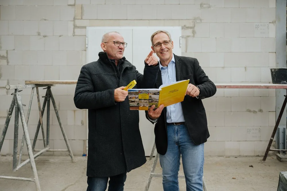 Zwei Männer in einem Baucontainer, einer zeigt auf ein Merkblatt, beide lachen und unterhalten sich.