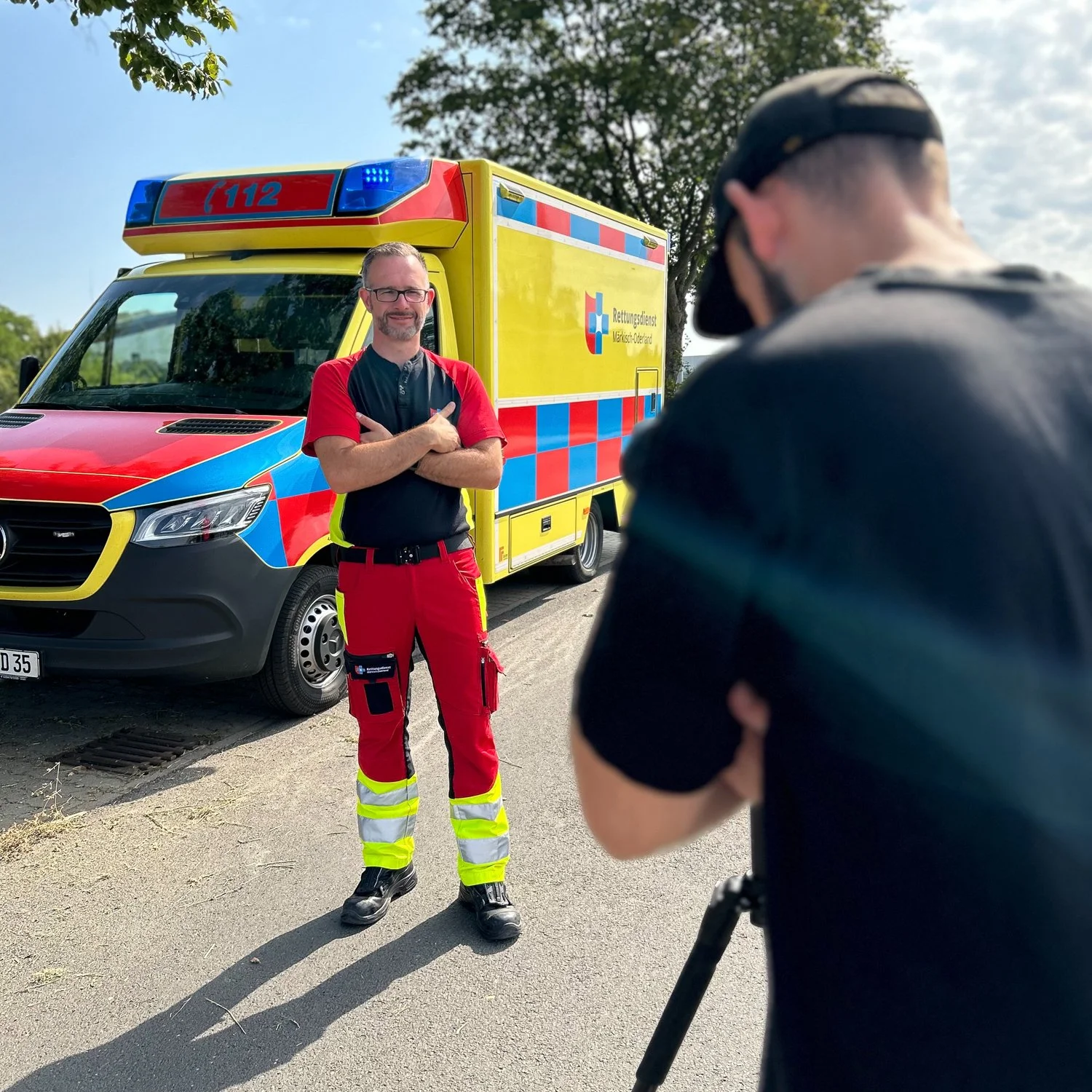 Ein Rettungssanitäter steht vor einem Rettungswagen mit verschränkten Armen, während ein Fotograf ihn fotografiert. Der Rettungswagen ist yellow mit rotem und blauem Melder. Es ist ein sonniger Tag mit einigen Wolken am Himmel.