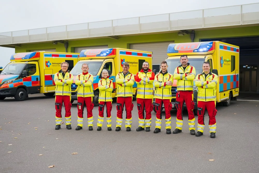 Eine Gruppe von acht Rettungsdienstmitarbeitern in gelben Warnwesten und roten Hosen stehen nebeneinander vor Rettungswagen.