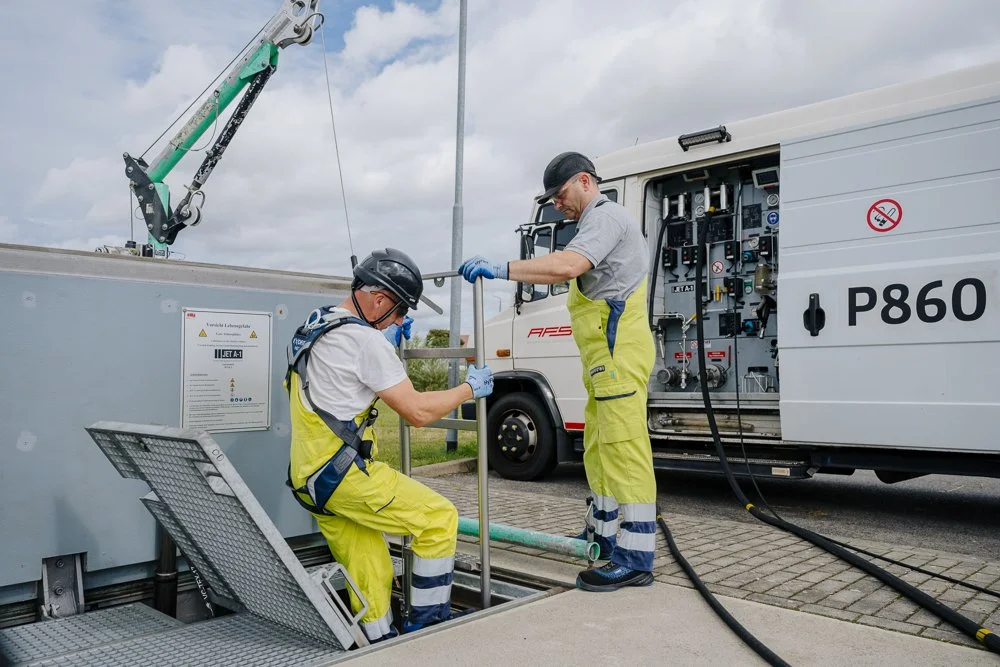Zwei Arbeiter in Schutzkleidung und Helmen arbeiten an einer technischen Anlage im Freien, eine im Hintergrund, eine im Vordergrund, mit einem Transporter im Hintergrund.