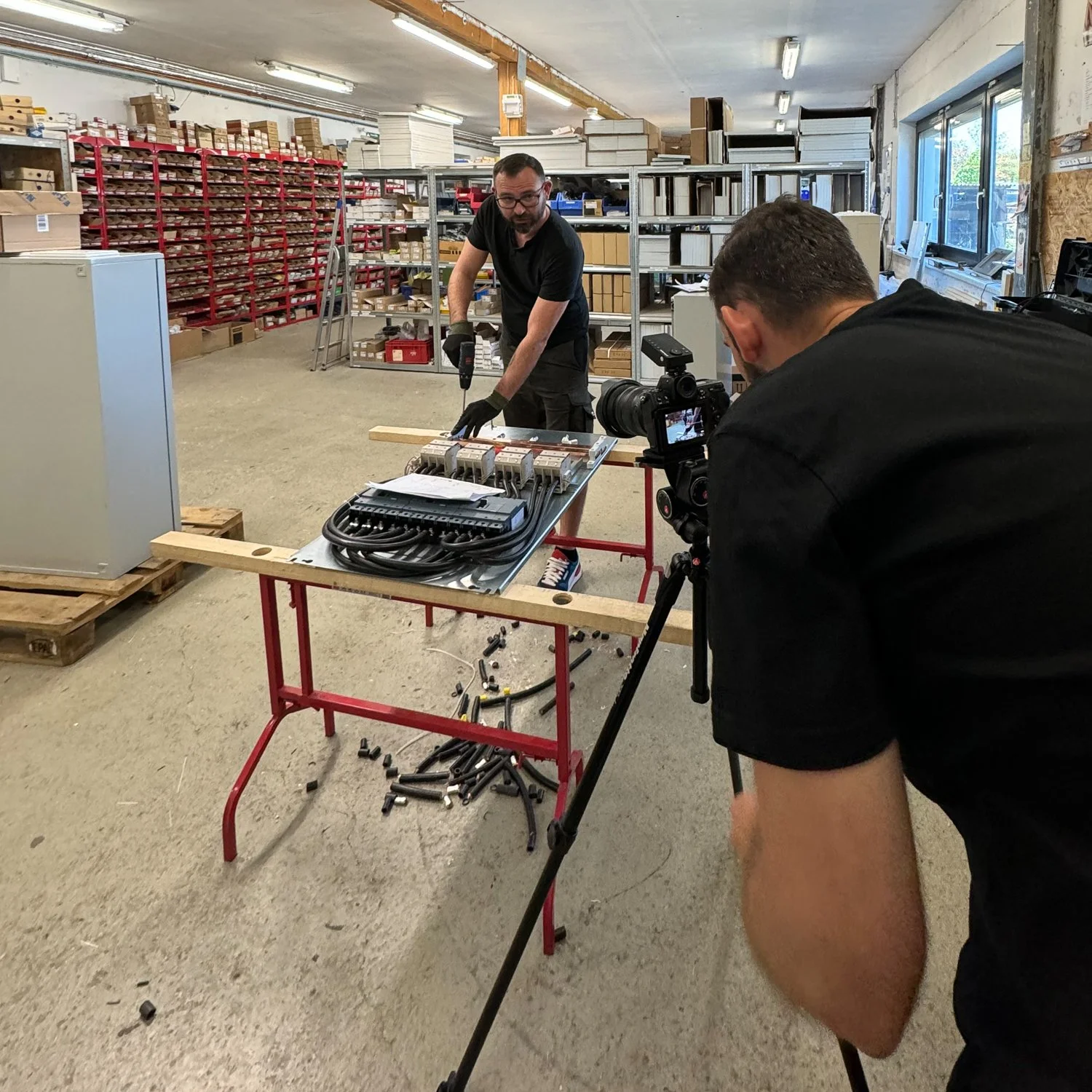 Mann installiert elektrische Komponenten an einem Arbeitstisch in einer Lagerhalle, während ein Videograf die Aufnahme macht.