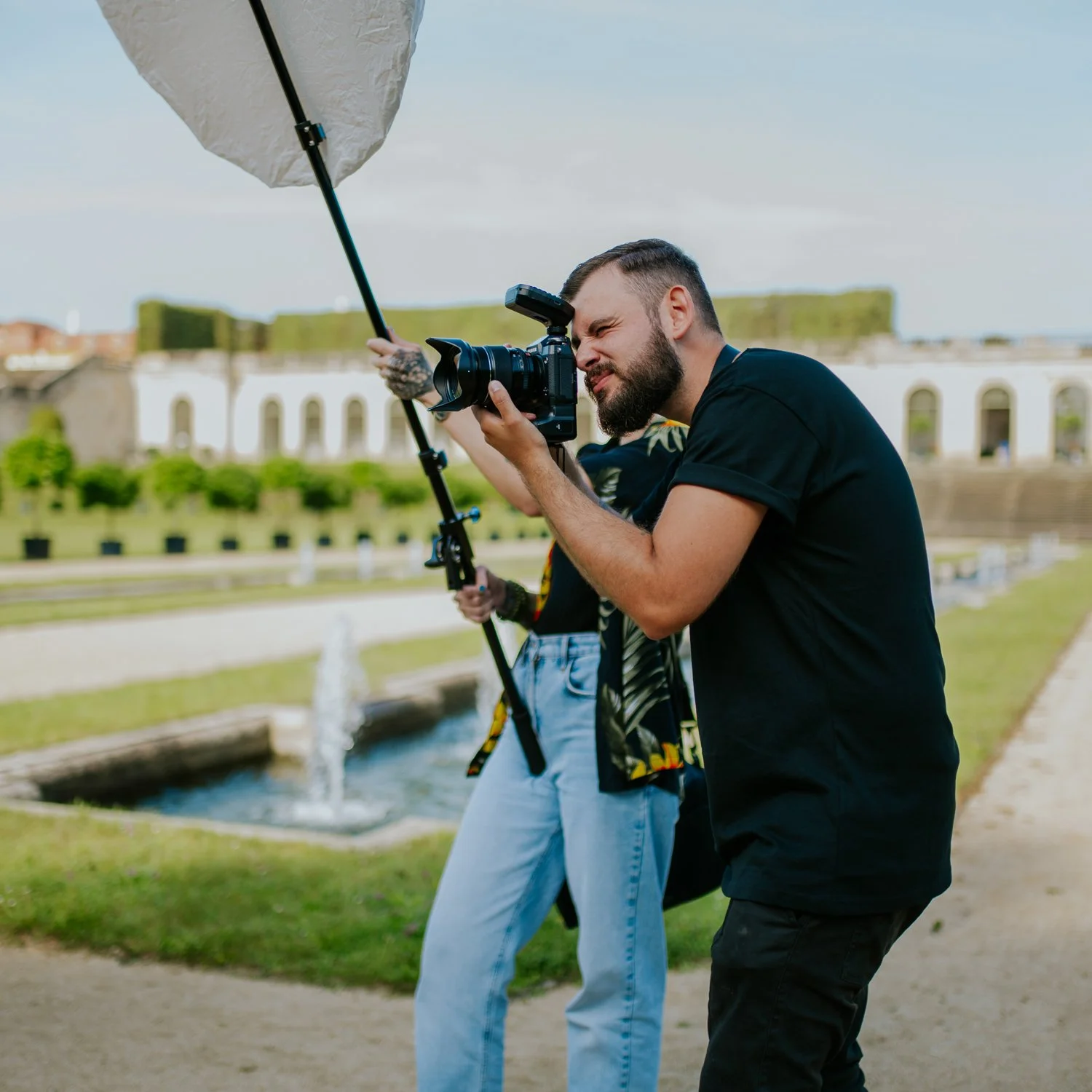 Zwei Fotografen beim Fotoshooting im Freien, einer hält eine Kamera mit einem Blitzaufsatz, im Hintergrund ein Wasserbecken und historische Gebäude.