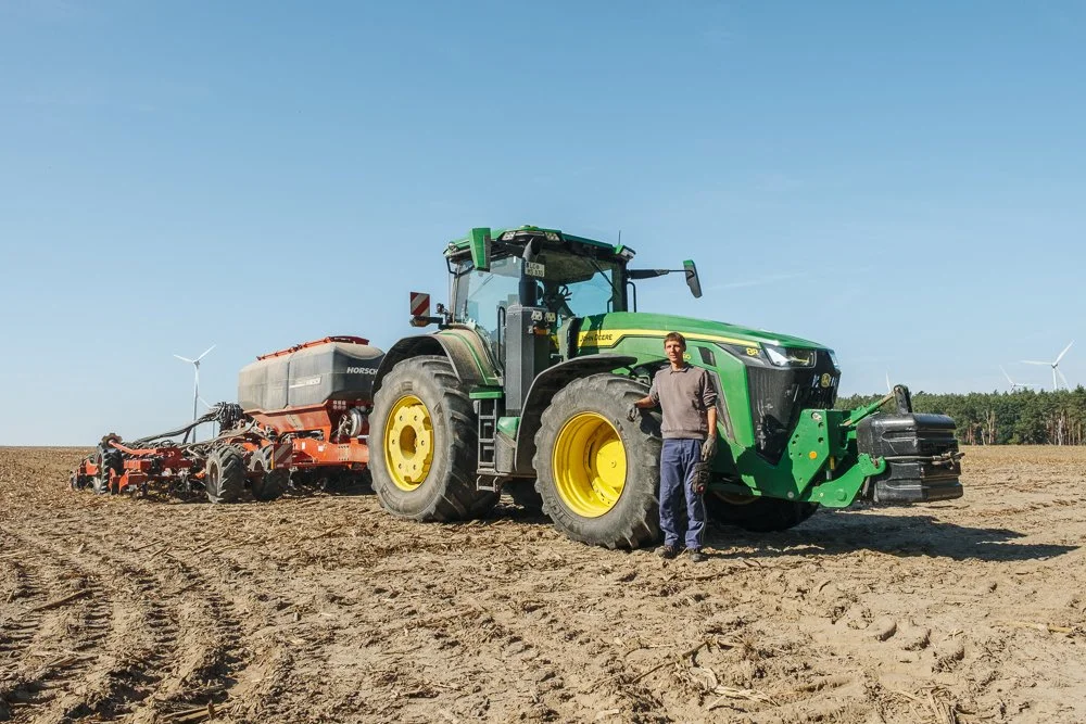Mann steht neben einem großen grünen Traktor mit gelben Rädern auf einem Feld, im Hintergrund sind Windkraftanlagen zu sehen.