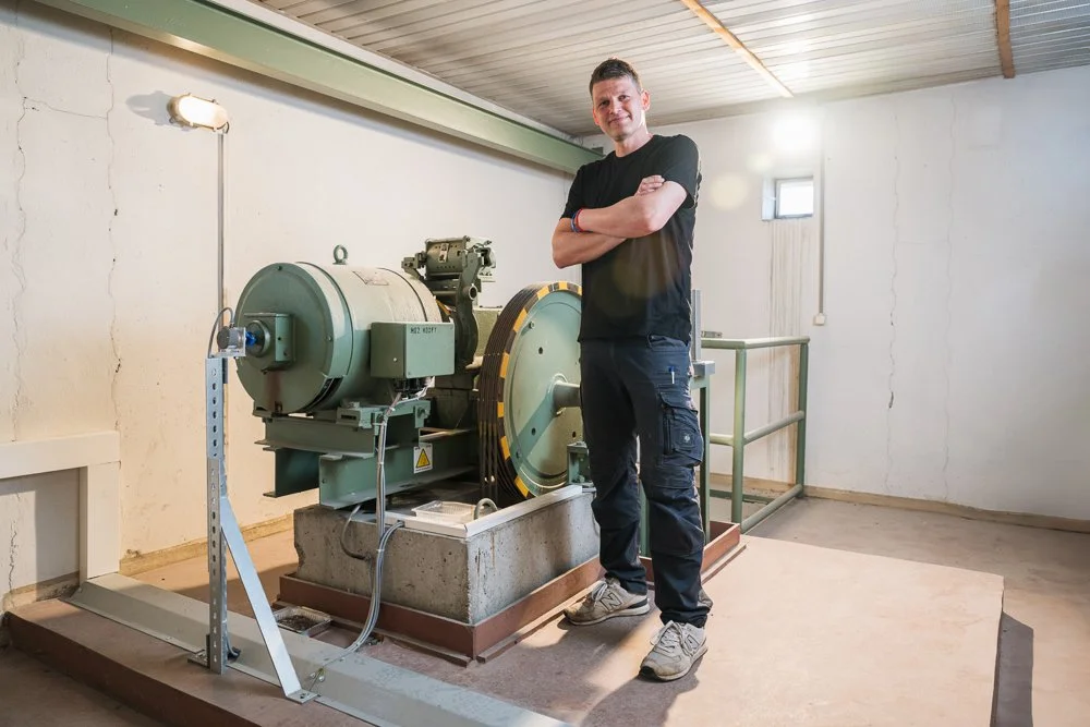 Ein junger Mann steht in einem Raum mit einer großen technischen Maschine, wahrscheinlich eine Zentrifuge oder eine ähnliche Vorrichtung, die auf einem stabilen Sockel montiert ist. Der Raum hat eine einfache, etwas ungepflegte Einrichtung mit uneben