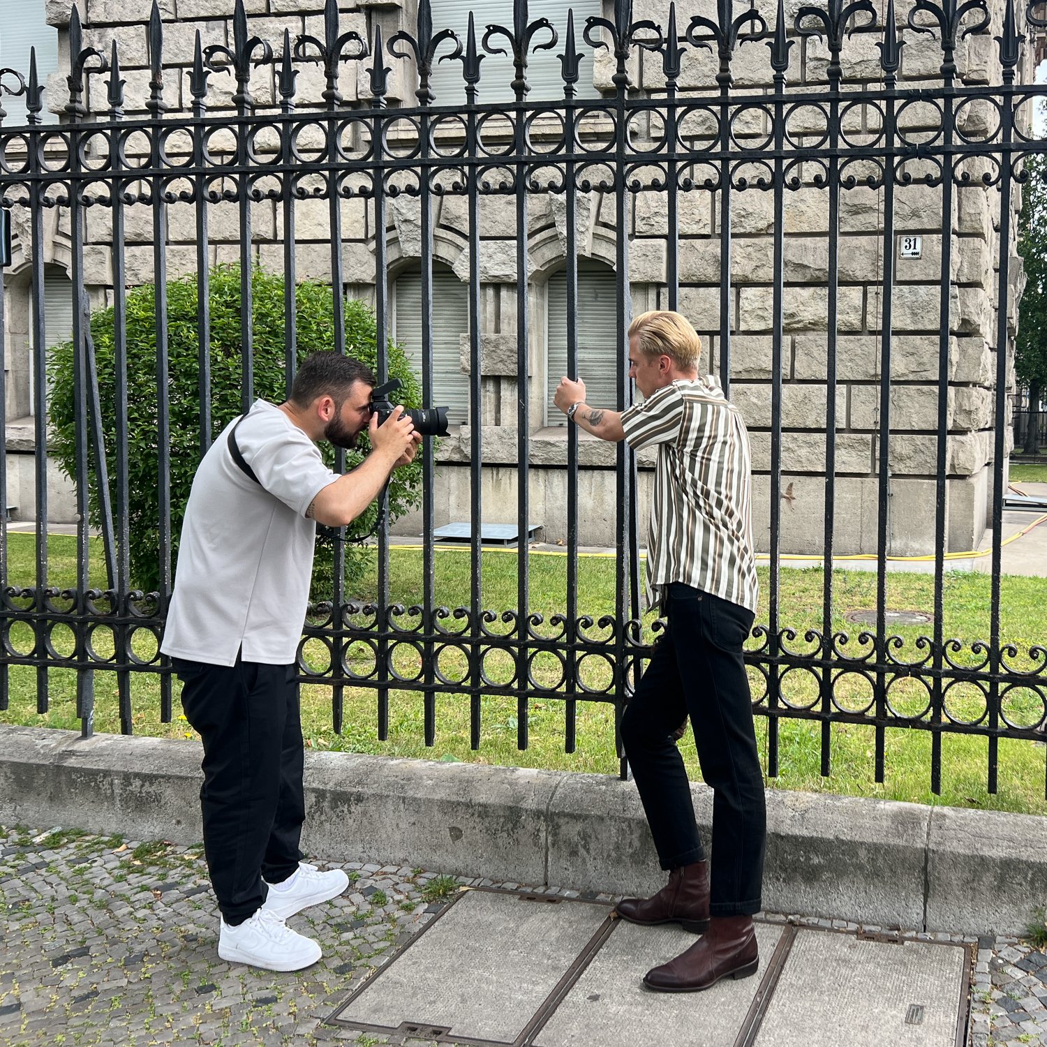 Zwei Männer fotografieren sich gegenseitig vor einem Schmiedegitter an einer städtischen Straße.