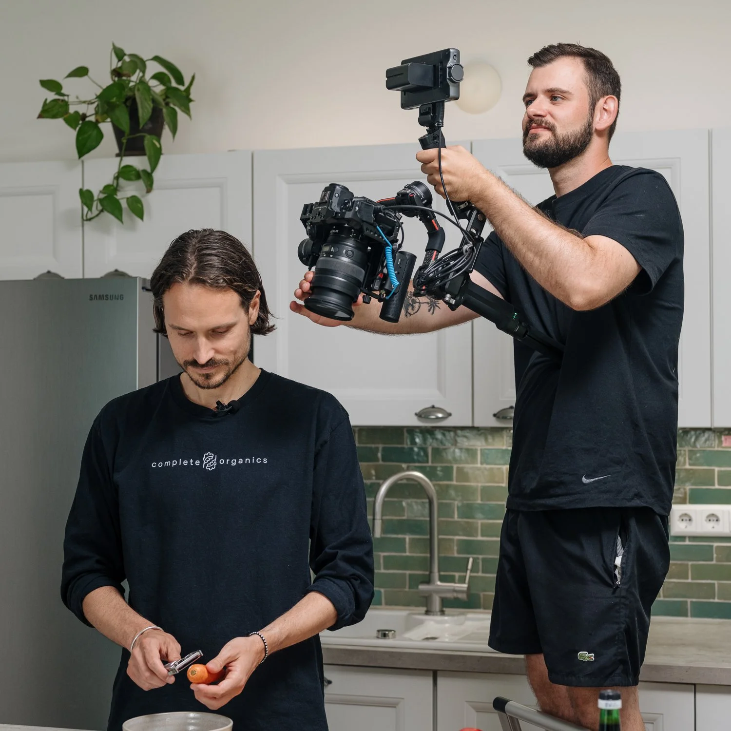 Zwei Männer filmen eine Frau, die ein Messer hält und eine Karotte schält, in einer Küche mit weißen Schränken und grünen Kacheln.