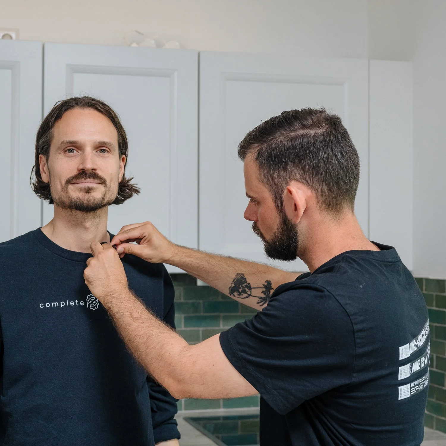 Zwei Männer in einer Küche, einer steckt dem anderen ein Kärtchen an die Kleidung.