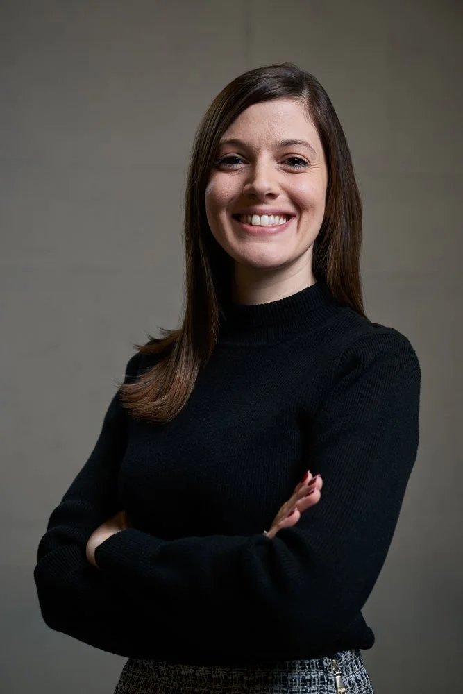 Eine lächelnde Frau mit braunen Haaren, die schwarze Kleidung trägt, steht vor einem grauen Hintergrund.