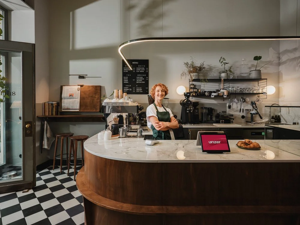 Lächelnde Barista in einem modernen Café hinter dem Barcounter mit Coffee-Maschinen, Pflanzen und einem digitalen Bestellterminal.