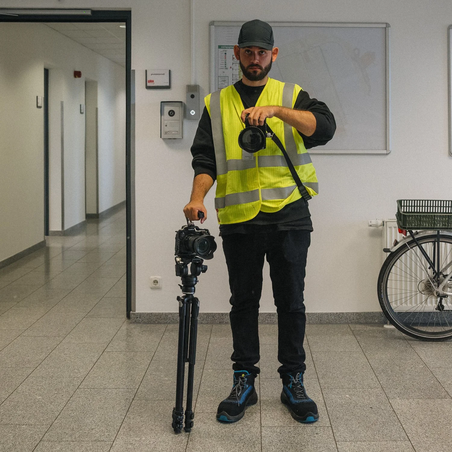 Mann in Gelber Sicherheitsweste hält Kamera und Stativ in einer Innenaufnahme.