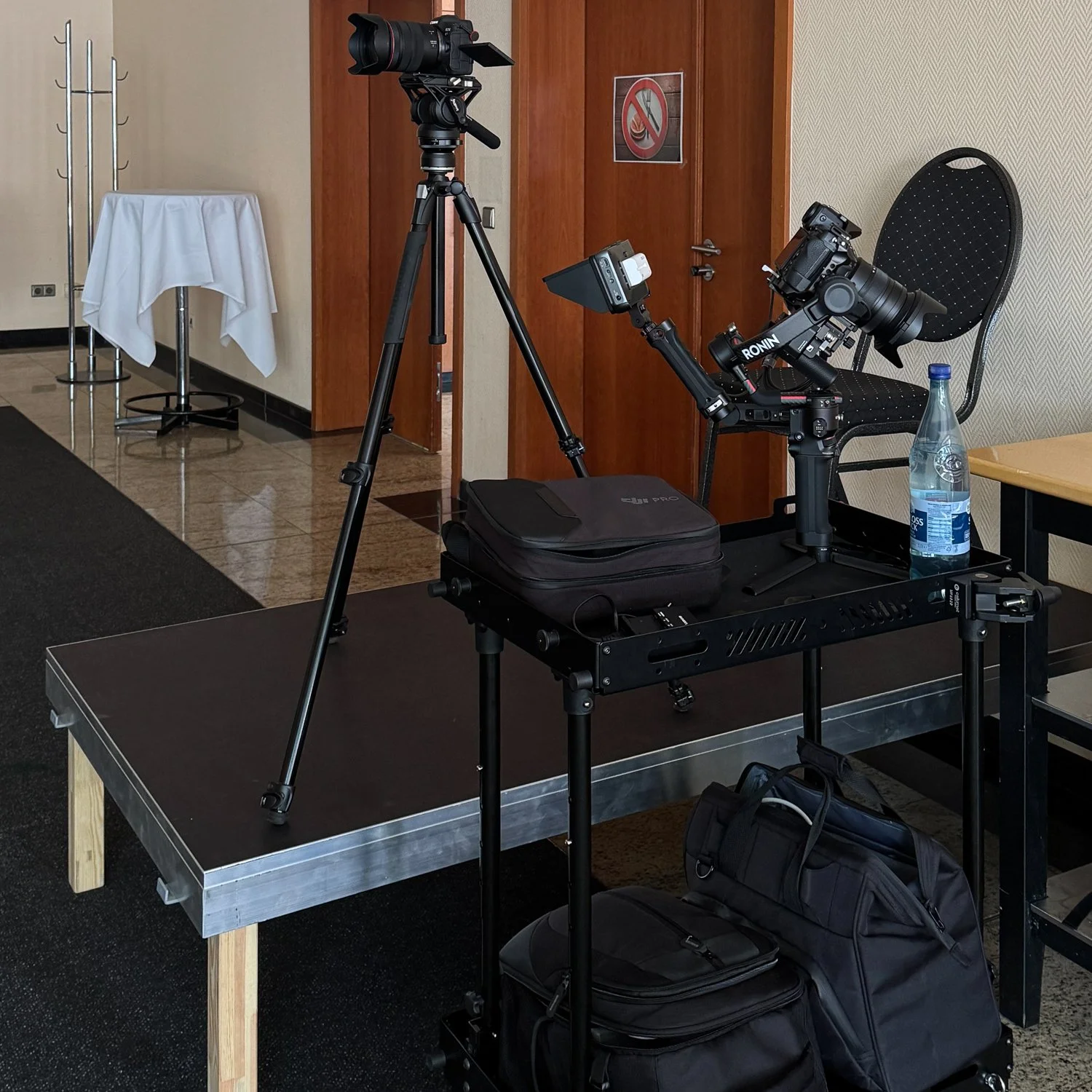 Fotografie einer Kamera- und Drohnenausrüstung auf einem Tisch in einem Raum mit Holzverkleidung und einer schwarzen Teppichboden. Es gibt einen Wasserflasche, eine schwarze Tasche und ein Stativ mit Kamera. Im Hintergrund sind ein Tablett mit weißer