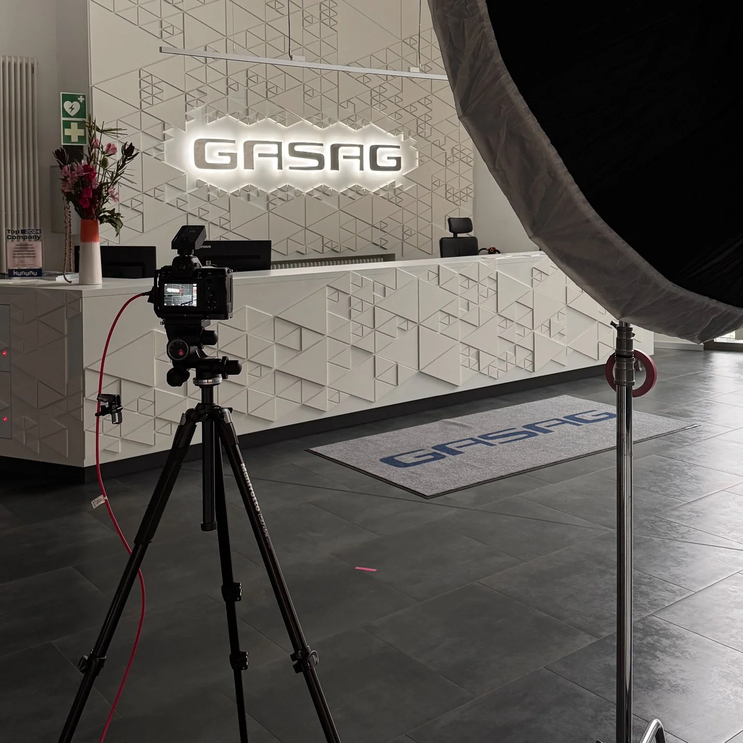 Aufnahmebereich mit Kamera vor einem weißen Empfangstresen mit geometrischem Muster, im Hintergrund ein beleuchtetes Logo 'GASAG' an der Wand, auf dem Boden ein Fußmatte mit dem Logo, links eine Vase mit Blumen, rechts ein Fotolicht mit Schirm.