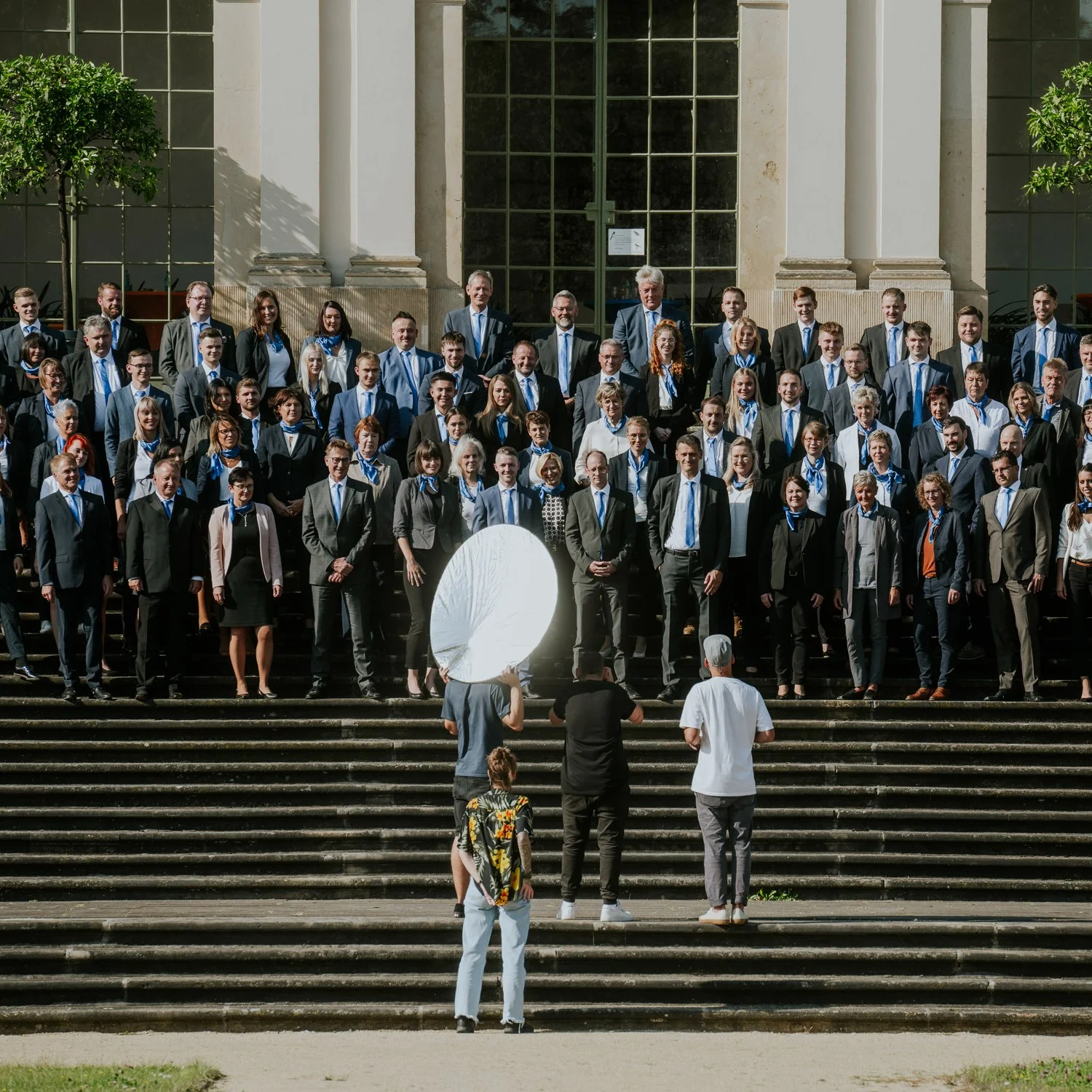 Gruppe von Menschen in formeller Kleidung auf den Stufen eines Gebäudes, wahrscheinlich bei einer Veranstaltung oder einem Gruppenfoto. Einige Fotografen fotografieren die Gruppe, mit Blitzlicht.