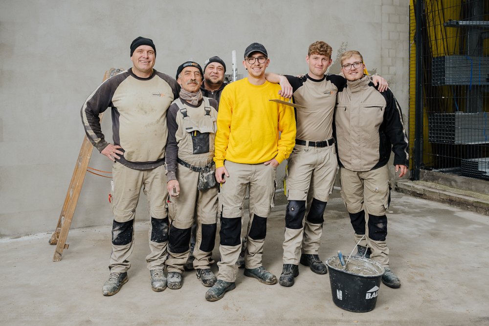 Sieben Junge Männer in Arbeitshosen und Jacken stehen zusammen auf einer Baustelle, manche mit Werkzeugen, vor einer grauen Wand.