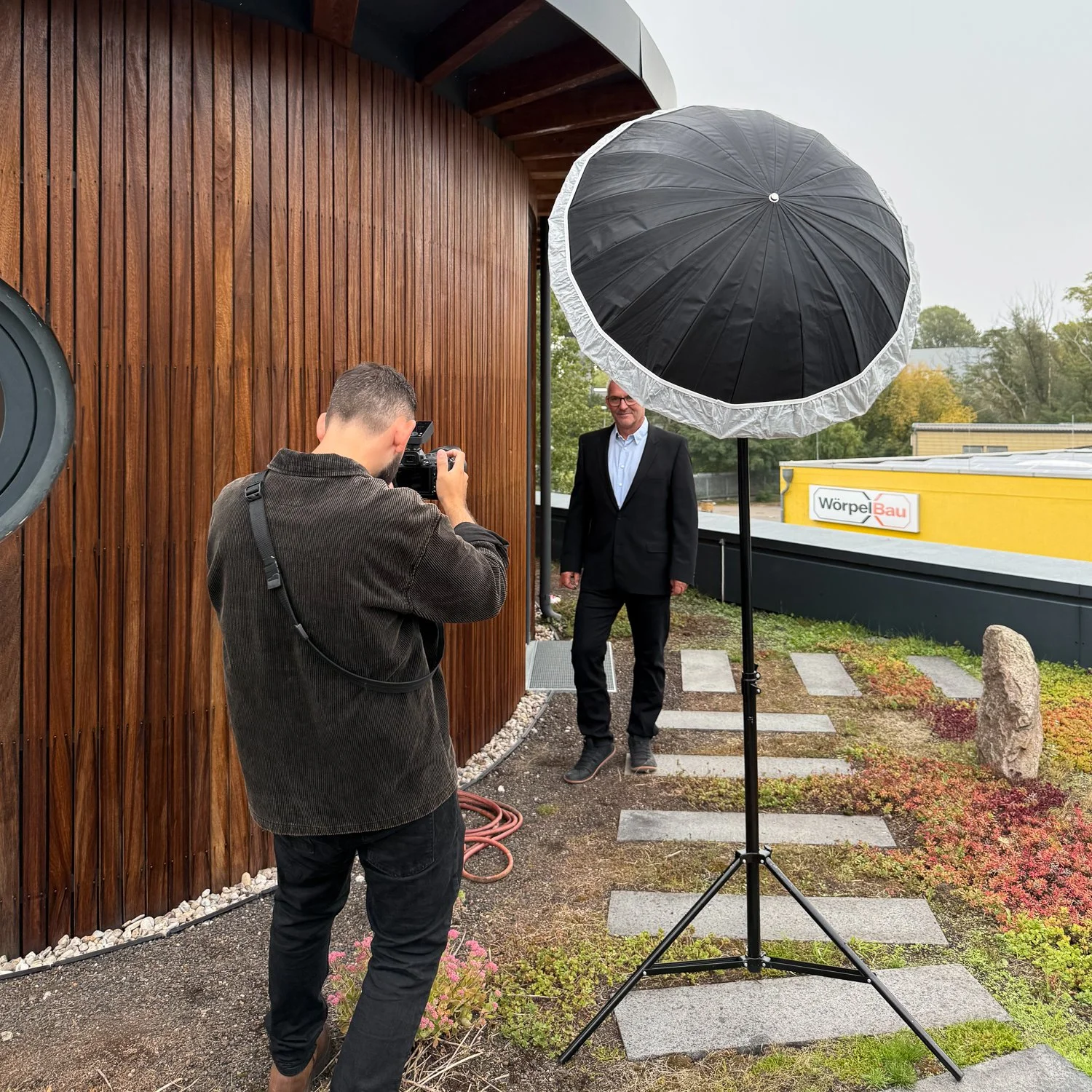 Ein Mann in Anzug posiert vor einer Holzwand während eines Fotoshootings mit einem Fotografen, einem aufstellbaren Studio-Blitz und einem schwarzen und weißen Schirm. Draußen, vor einem Gebäude mit einem Schild, das 'Wörpel Bau' zeigt, sind Bäume und