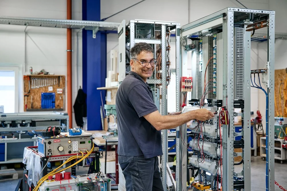 Ein Mann in einer Werkstatt arbeitet an einer großen elektrischen Schalttafel, umgeben von Werkzeugen und technischen Geräten.