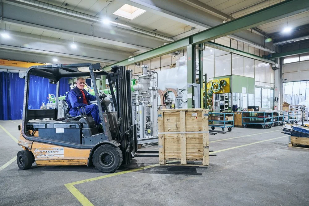 Ein Mann fährt mit einem Gabelstapler in einer Fabrikhalle, umgeben von Metallregalen, Werkzeugen und Verpackungsmaterialien.