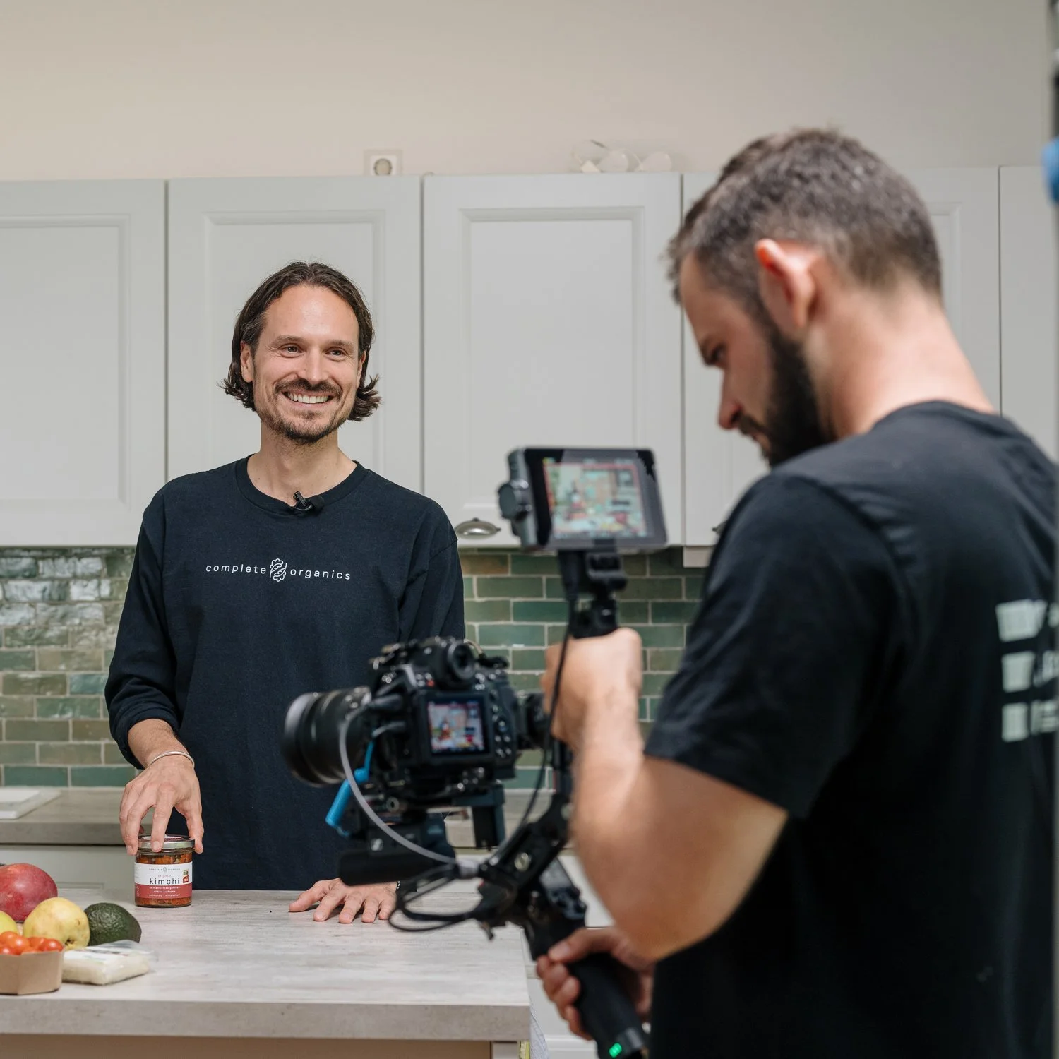 Ein Mann mit braunen, lockigen Haaren, trägt ein schwarzes T-Shirt mit dem Logo 'complete organics' und steht in einer Küche. Er lächelt und hält ein Glas. Im Hintergrund sind weiße Küchenhängecabinetts und eine grüne Fliesenspiegelung. Vor ihm steht