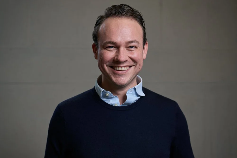 Lächelnder Mann mit dunkelblauem Pullover und Hemd vor grauer Wand.